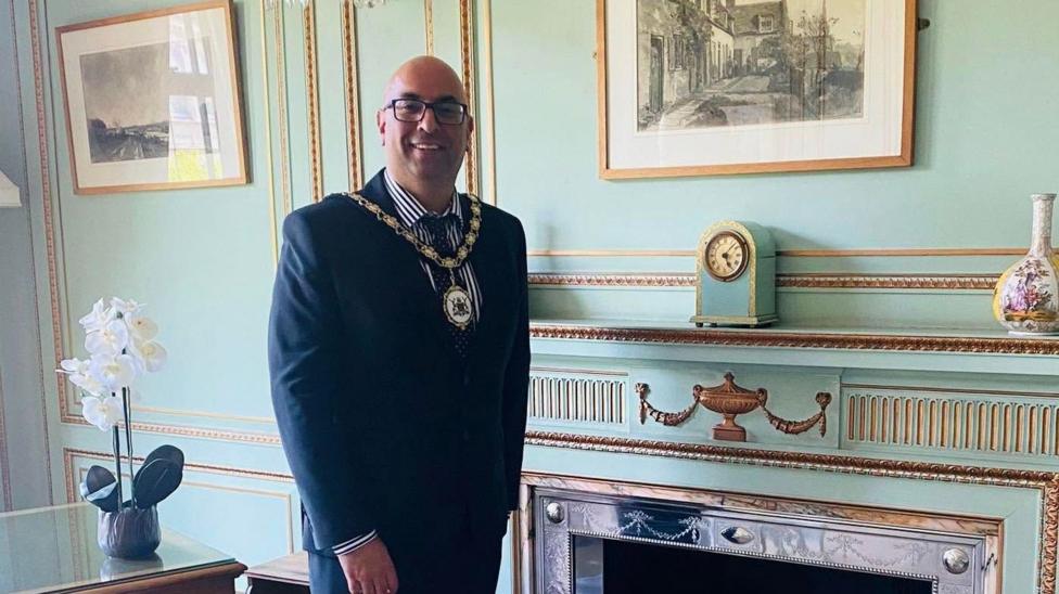 A man wearing ceremonial chains and a suit. He is stood in a room with blue walls