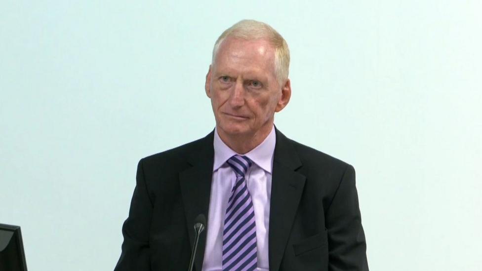 Michael Tonge giving evidence at the Omagh Bombing Inquiry. He has short blonde hair, wearing a black blazer, purple shirt and purple striped tie.