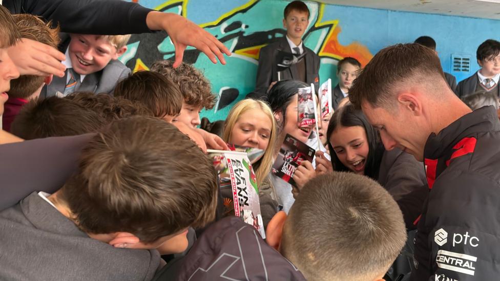 A man with brown hair wearing a black sports jacket and surrounde dby children in grey school uniforms, some holding flags with the name Elfyn Evans on it.