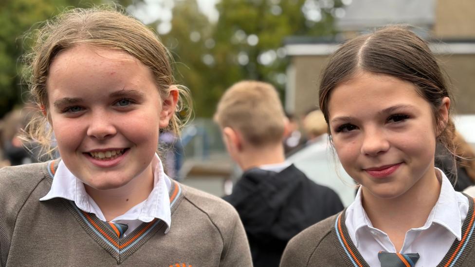 Two young girls, both with tied back hair, both wearing white shirts and brown jumpers, both looking at the camera and smiling.