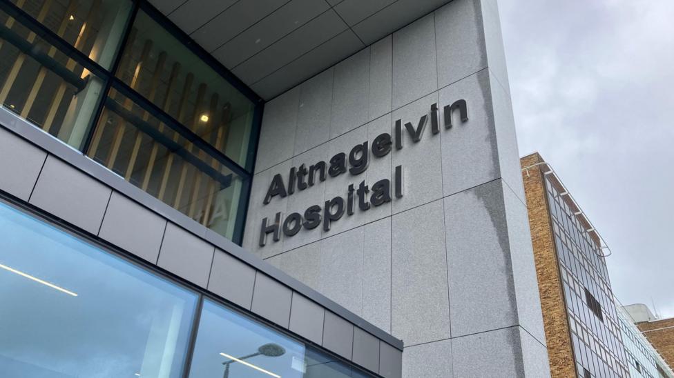 A close up of a sign outside Altnagelvin Hospital, which says the name of the building in large lettering. It is a tall building with a mix of facades including brown brick and grey cladding.