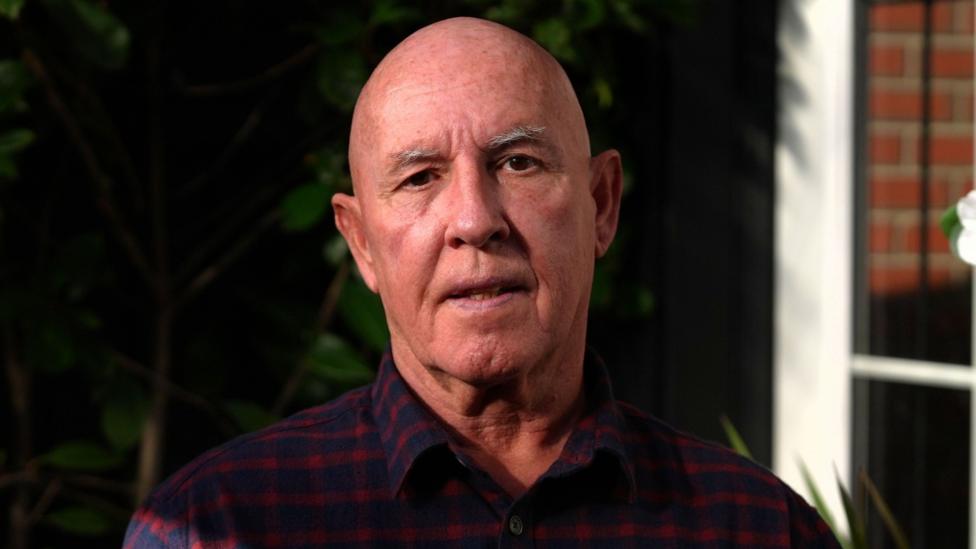 A man in a blue and red check shirt looks at the camera