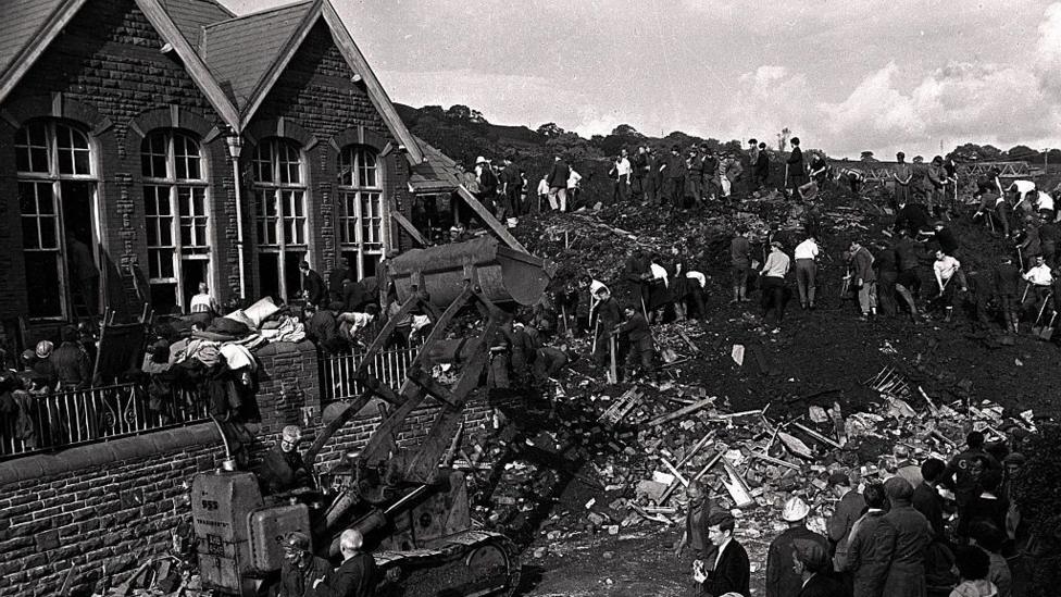A black and white photo of a digger working through the rubble, with people searching in the slurry