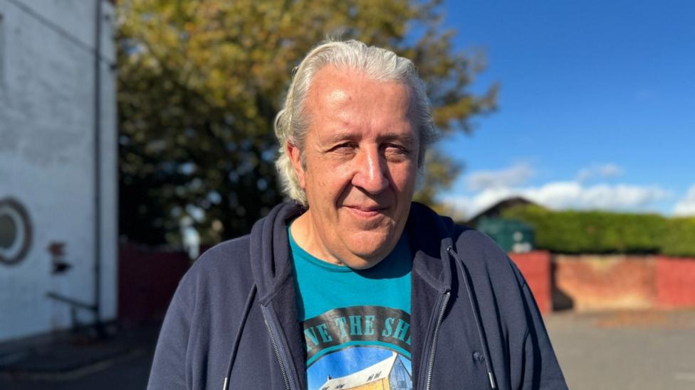 A head and shoulders photograph of Mark Newlands. He has white hair and is wearing a t-shirt and navy hoodie jacket.