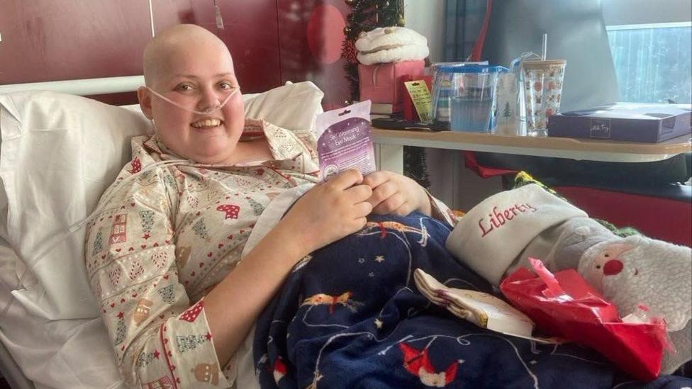 Liberty in a hospital bed intubated, wearing Christmas pyjamas. She has no hair due to chemotherapy treatment 