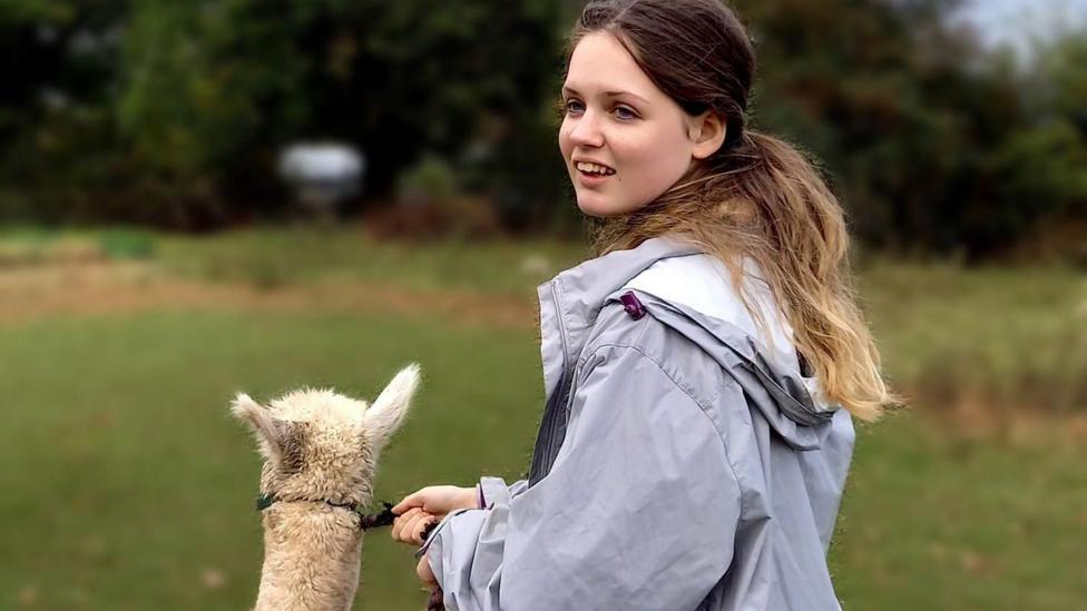Liberty walking an alpaca at a farm, wearing a long lilac waterproof jacket and her hair tied up
