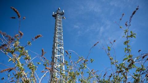 5g mobile phone mast on April 04, 2020 in Cardiff. The mast can be seen standing very tall amid blue skies. Some bushes can be seen at the forefront of the photo.