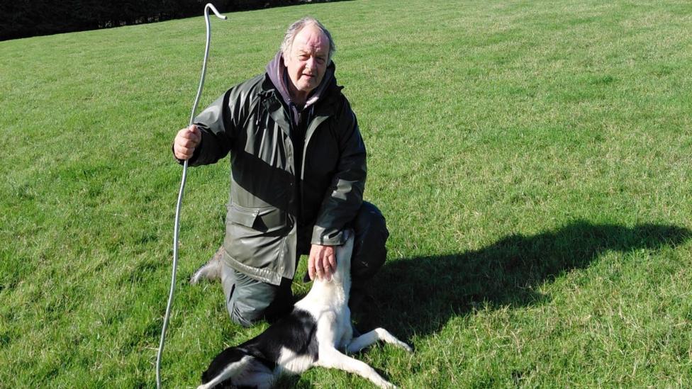 Alan Watson is knelt down on the grass. He holds a cane in his left hand and wears a dark green parka. He has silver short hair and looks directly at the camera. Green grass can be seen on the hill behind him, along with trees and a mast in the far background.