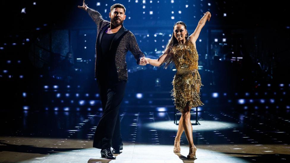 A couple on a dance floor during a routine where their arms are outstretched. The woman is wearing a gold knee length dress and the man wears black trousers and t-shirt with a pewter coloured jacket.There are lots of fairy lights in the background.