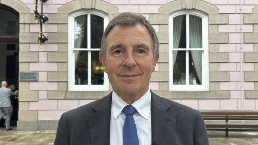 Alan Maclean pictures outside the staes building. He has short brown hair, greying at the temples. He is smiling. He is wearing a grey suit, white shirt and blue tie.