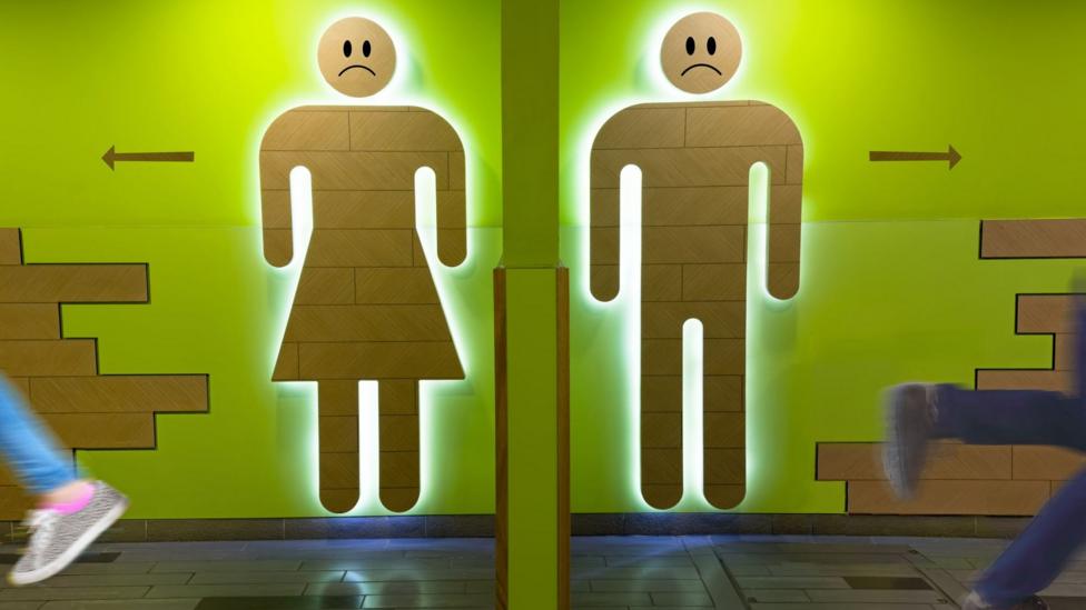 Wood-effect male and female toilet symbols are lit from behind on a bright green wall. A central pillar divides the two signs, while blurred passers-by move across the foreground.