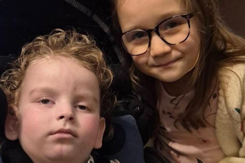 A young girl with glasses smiling for a photo next to her brother who's in a wheelchair