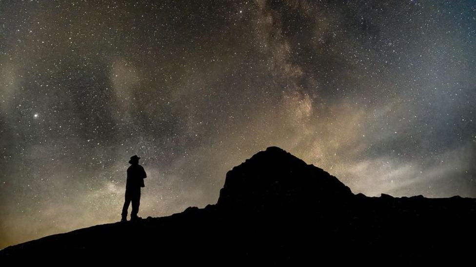 The sky is filled with stars and natural light, while a man stands in silhouette on a hill, with the peak also in silhouette.