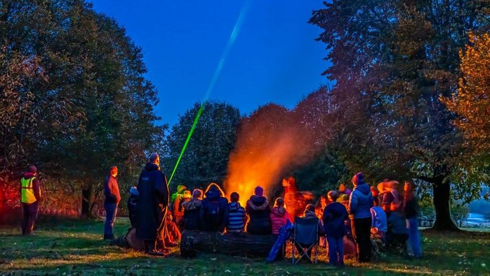 A group of people all sit in coats on logs and camping chairs around a smoking fire. One man at the back is pointing a green laser into the sky. The group, which is facing away from the camera, is surrounded by trees.