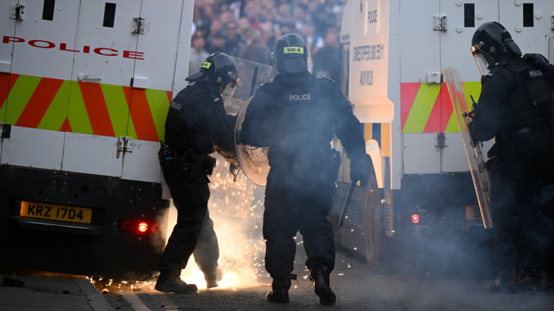 Three officers are in riot gear. The word police is stated on the back of their clothing. The are standing behind PSNI land rovers. A crowd is facing them. 
