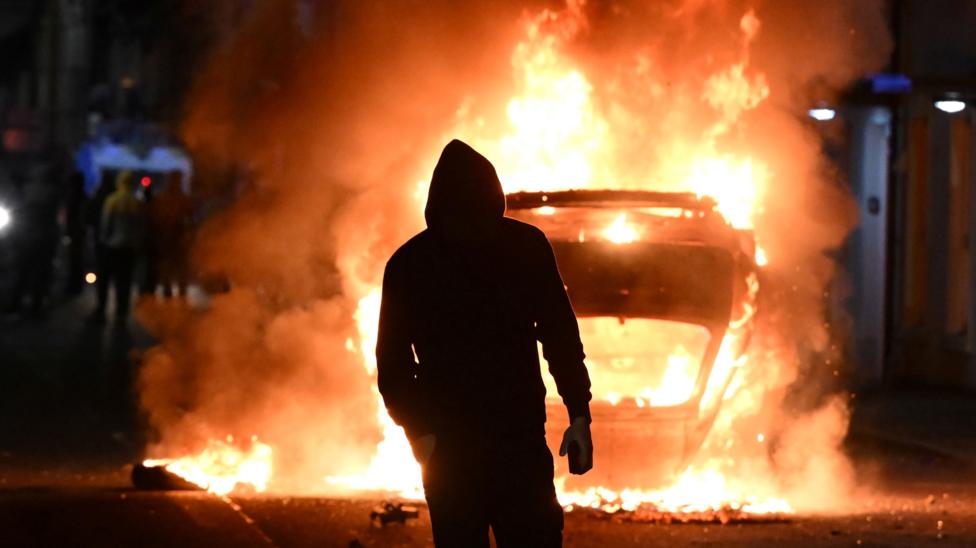 A hooded male is standing in front of a burning car. It's night. Other people are in the background. 