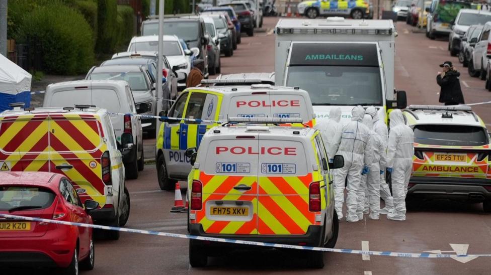 A Glasgow street, with a row of houses stretching into the distance. Several police vans, cars and an ambulance are parked along part of the street, with police tape sealing off part of the street an forensic officers in white overalls in the road. A blue police forensics tent has been set up on the pavement.