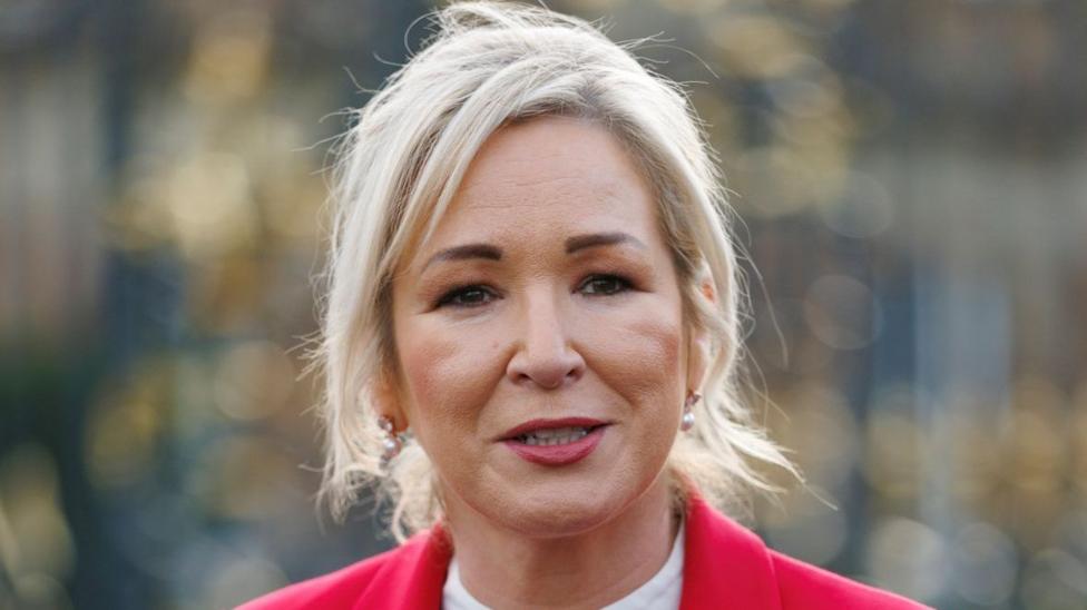 O'Neill has long blonde hair tied up wearing makeup, a white top and red blazer.