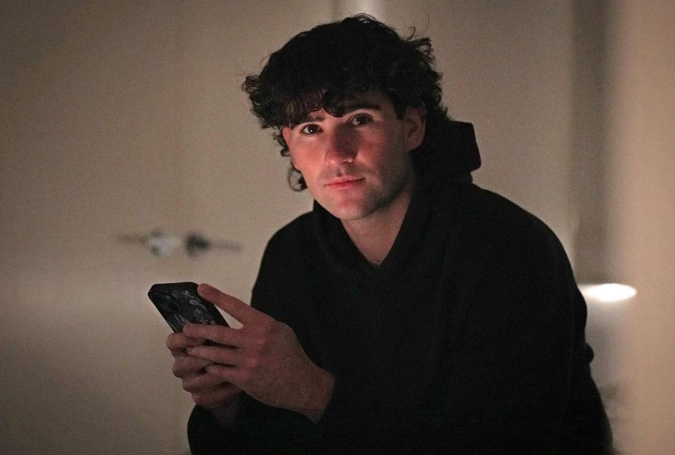 Flynn Martin, who has dark curly hair, looks at the camera while holding a mobile phone which lights up his face