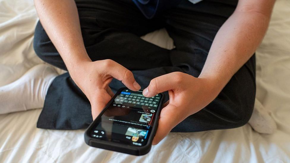 A teenage boy looks at an iPhone smartphone screen showing a messaging app in his bedroom