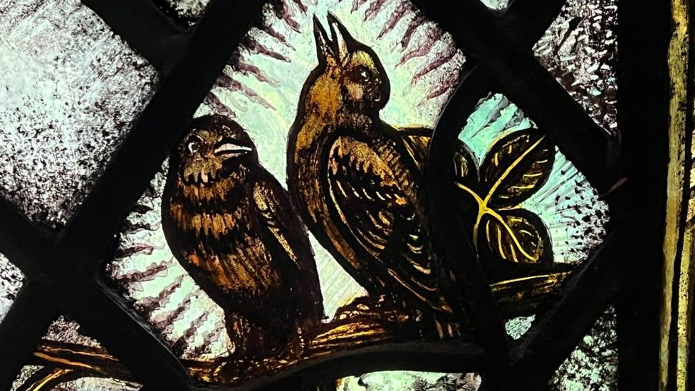 A yellow-toned stained glass window depicting two birds and a flower.
