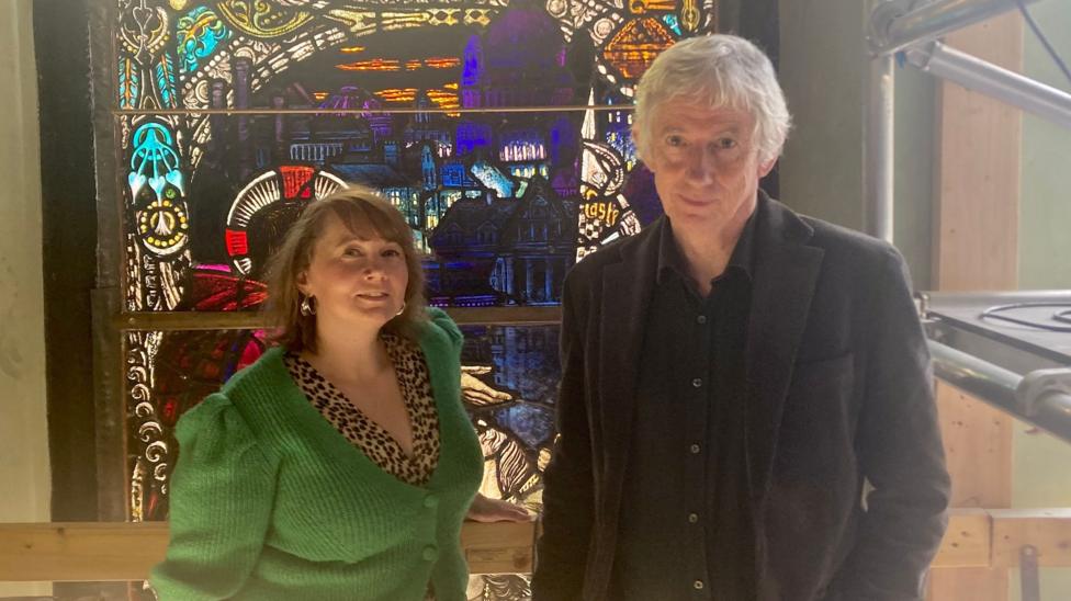 Two people stand beside a restored stained-glass panel inside a church being converted into a museum.