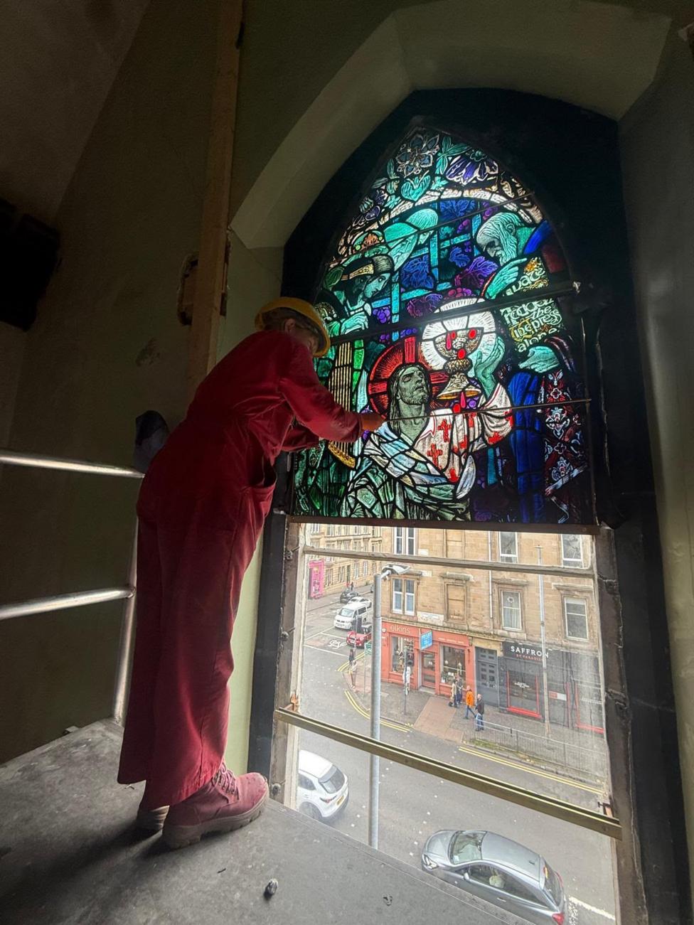 Conservator wearing red overalls and a helmet reinstalls a stained-glass window. Below you can see a city street.