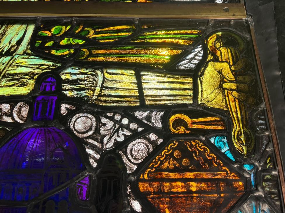 A stained glass window showing an arm painted in yellow holding a staff, other parts of the art show a purple church building.