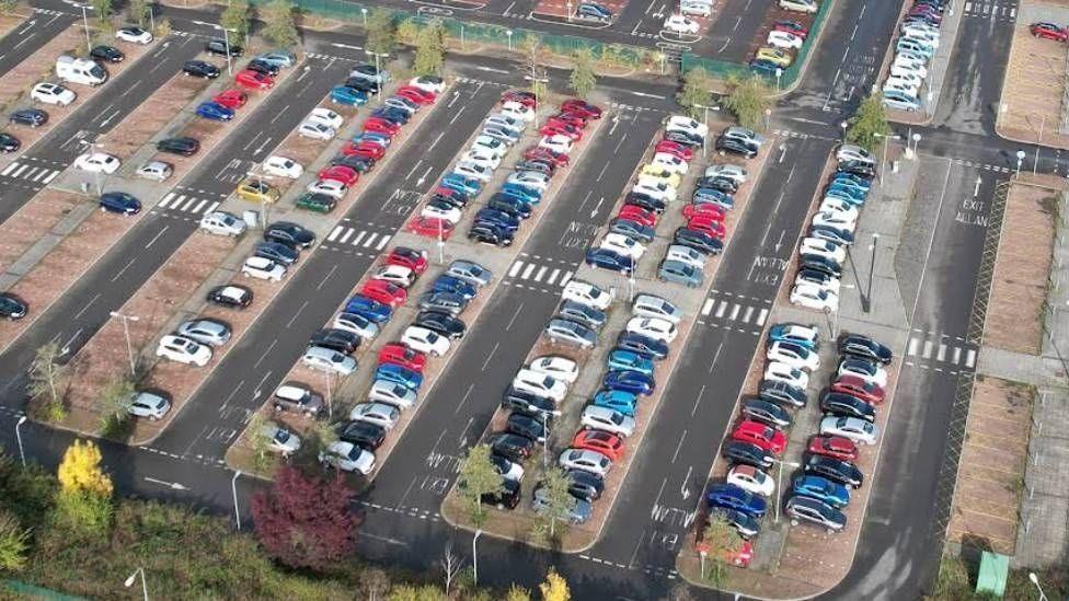 A picture of a car park taken from the air. Cars and lined up in a row with roads and trees in the middle.