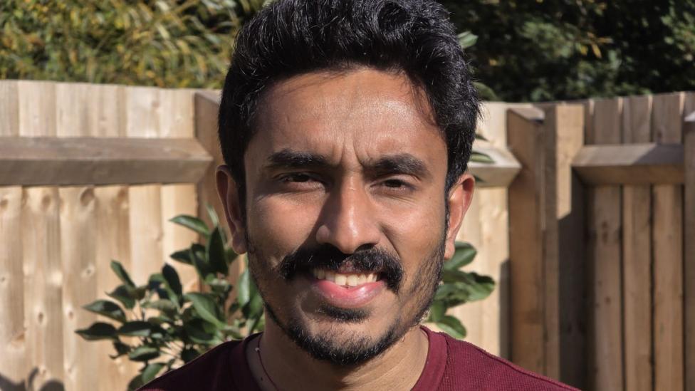 A close-up shot of a man wearing a red tee shirt with fencing and a tree behind him.