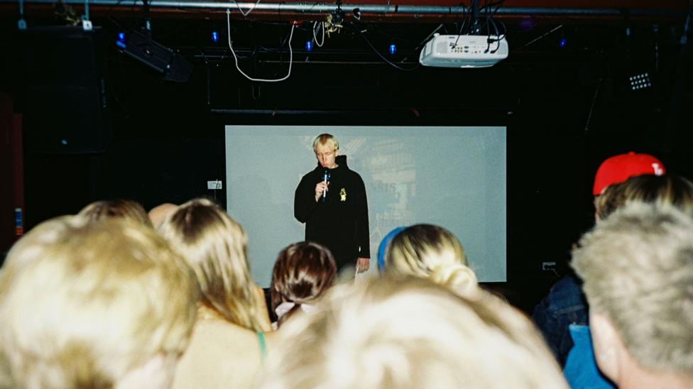 A man stands with a microphone in front of a projector screen speaking to the crowd.