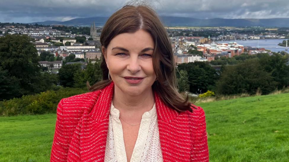 Sinead McLaughlin is wearing a red jacket and white top. She has long cbrown hair, and is pictured standing iin a field , with a cityscape behind her.