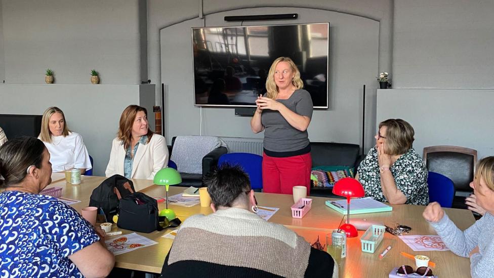 A group of women sit around a table listening to Leigh Williams speak. Leigh is standing up, gesturing with her hands. She is wearing red trousers and a grey top and has blonde, shoulder-length hair. On the table there are colourful lamps and colouring-in sheets.