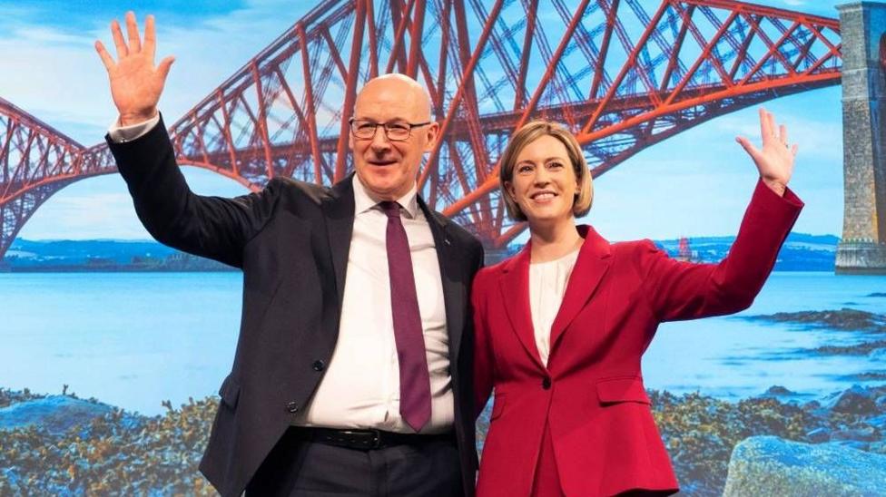 John Swinney wearing a darks suit and purple tie waving his left arm. His other arm is around Jenny Gilruth, who is wearing a red suit. She also has her arm outstretched. Behind them is a picture showing the Forth Rail Bridge.