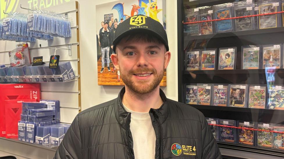 A man with ginger hair and a ginger beard, wearing a black baseball cap, a beige T-shirt and a black jacket.