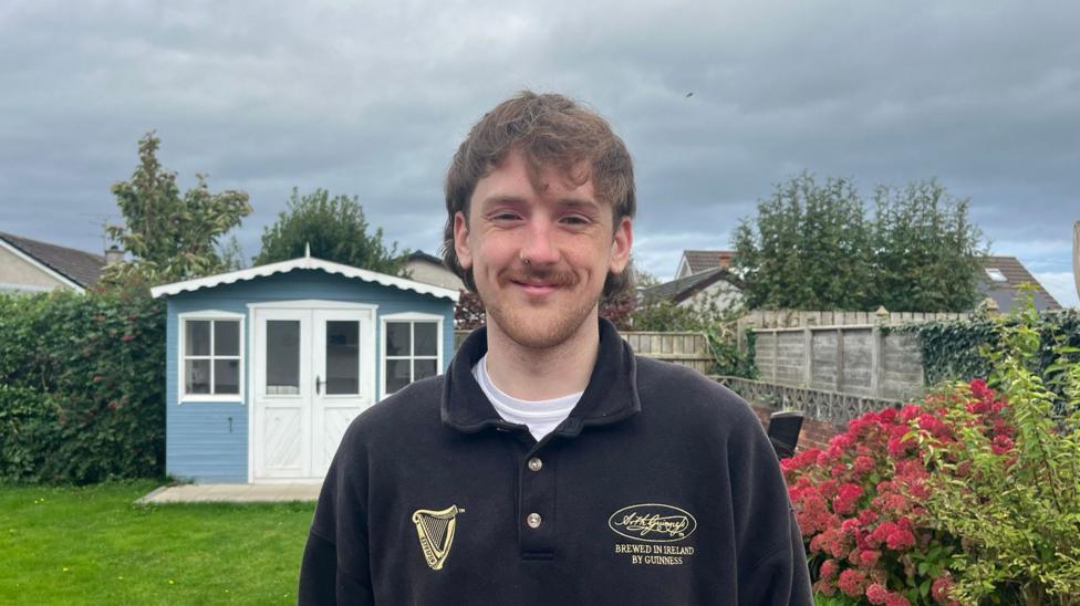 A man with brown hair, a brown moustache, wearing a black and gold fleece jacket, standing infront of a garden and a blue shed.