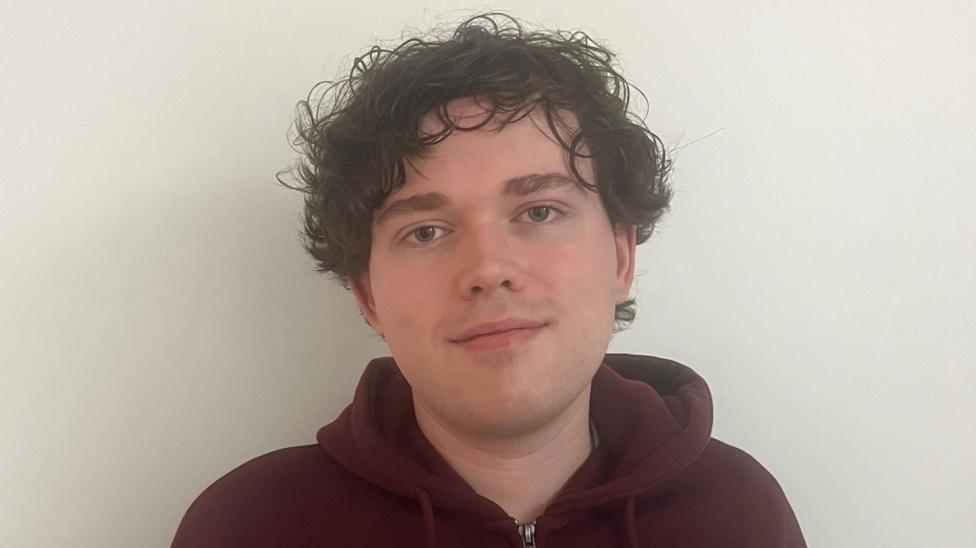 A young male with a maroon hoodie on. He has curly dark hair and is standing infront of a white background