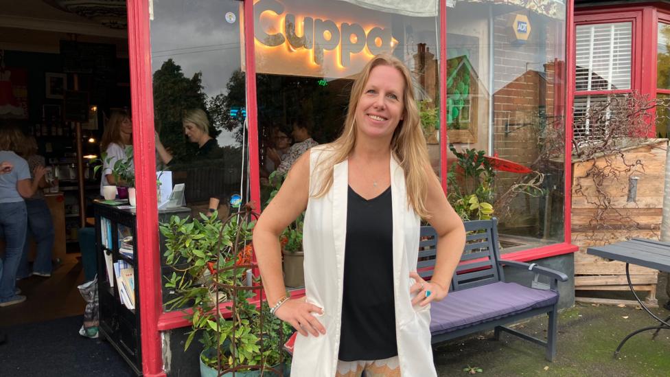 Vanessa Fullerton-Batten stands in front of a coffee shop with her hands on her hips. She has blonde hair and is wearing a black vest with a sleeveless white jacket. 