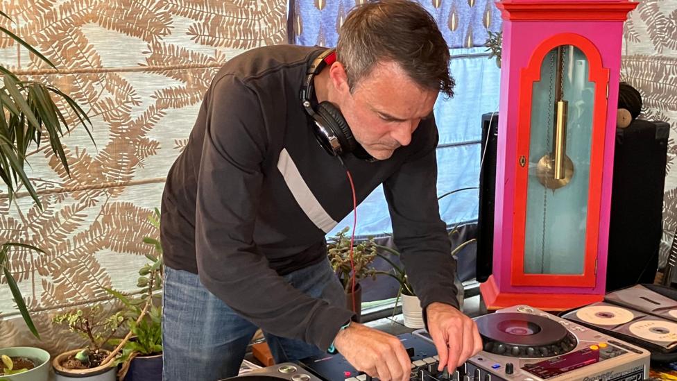 Ben Kay wears a black top and has headphones around his neck, and is DJing on some decks. 