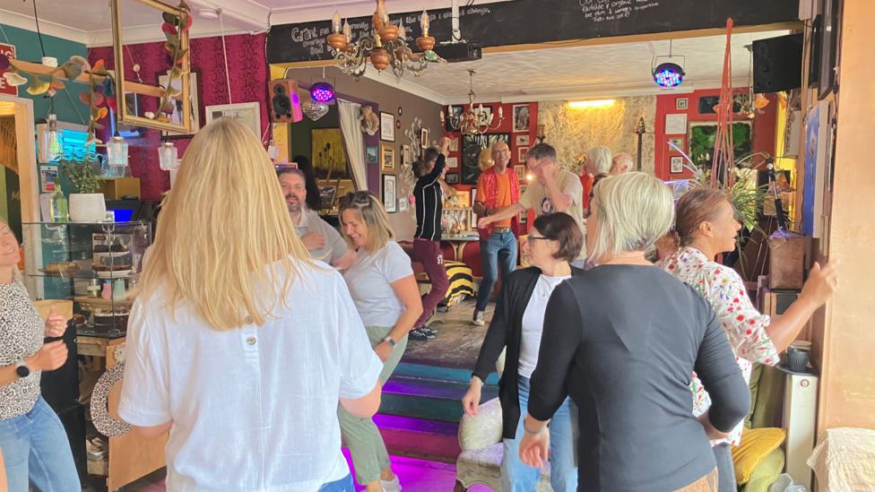 A group of people are dancing in a cafe. There is a chandelier on the ceiling and a chalkboard on the wall. 