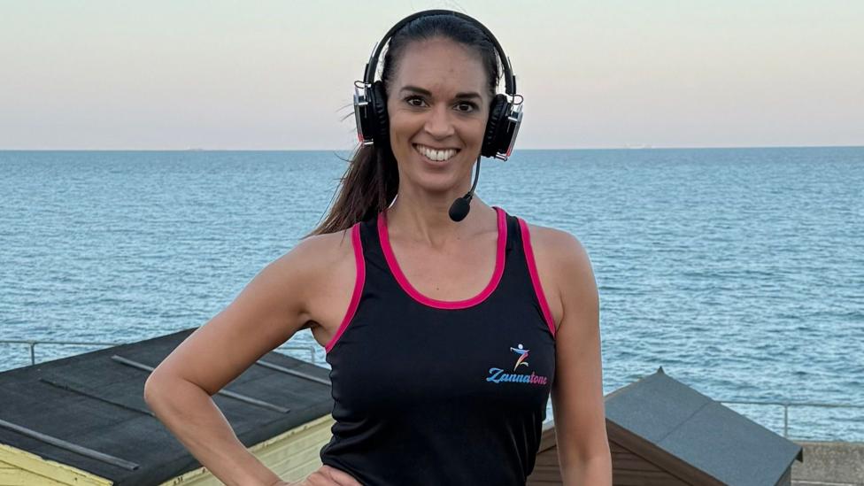 Susannah Hinchey stands with her hand on her hip in front of the sea and some beach hut roofs. She is wearing headphones, with a microphone, and a fitness vest. 