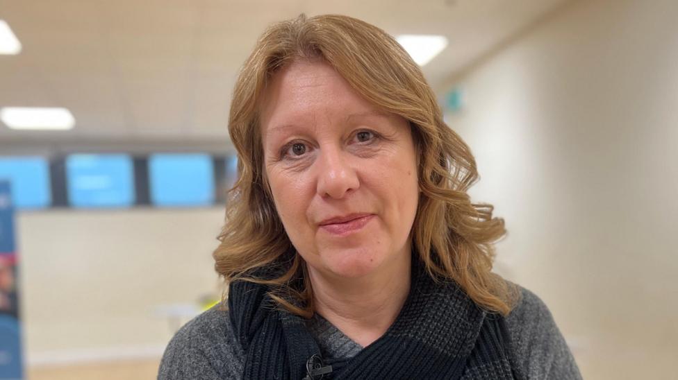 Tracey Taylor is in the middle of the image. She has light brown shoulder length hair and is wearing a grey jumper with a black scarf. She is inside a well-lit room.