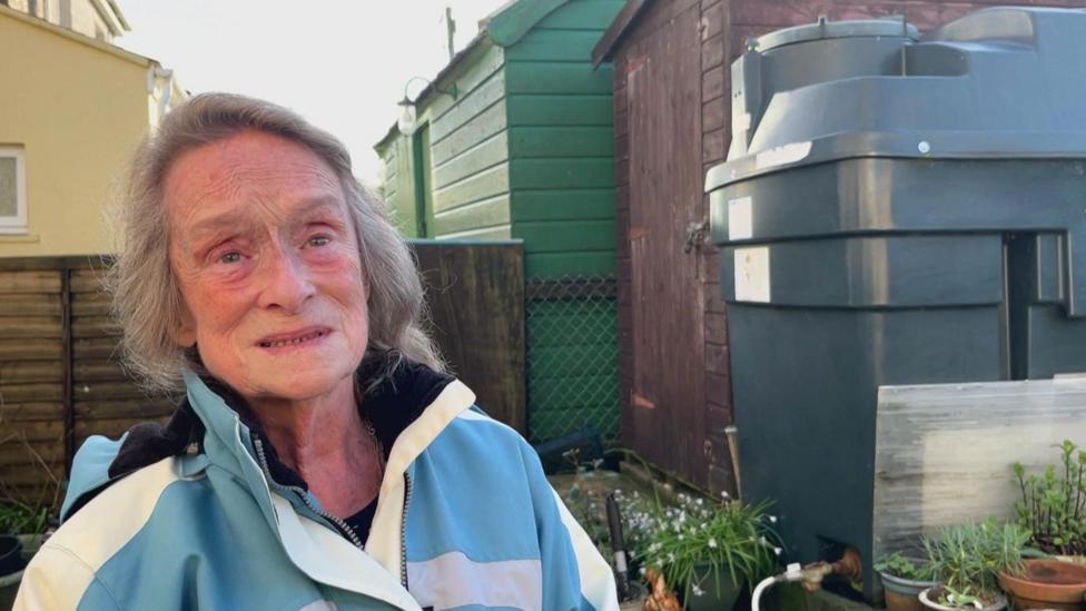 Pauline Trubody stands in her garden next to a dark green heating oil tank, which is next to a brown shed. She has long grey hair and is wearing a blue and white coat.