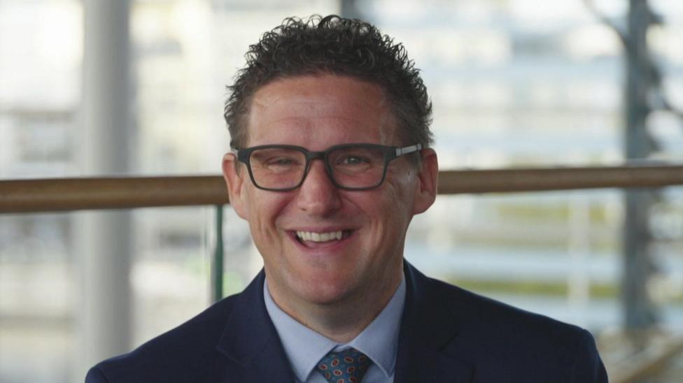 Jason O'Connell, a middle aged man with short brown hair, glasses, and a navy suit and blue tie, smiles.