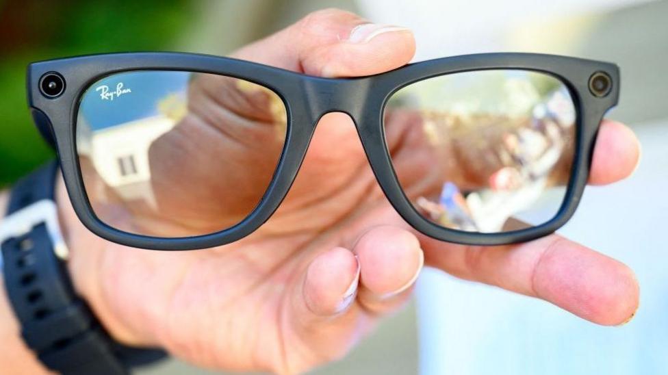 A pair of Meta glasses, being held in someone's left hand. They are black with a camera visible on each side. The person is wearing a watch.