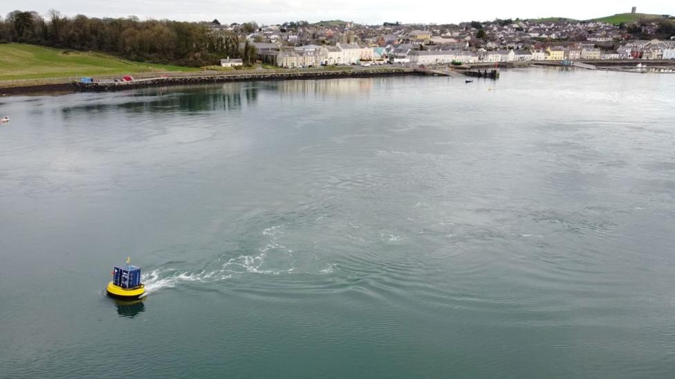 Aerial view of a floating marine energy device operating in a sheltered harbour, leaving a wake on calm water with a coastal town and shoreline visible in the background.