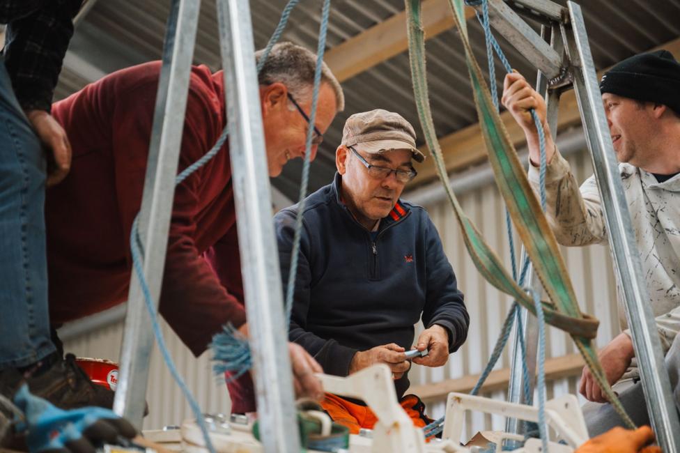 Workers assemble or inspect equipment within a metal frame structure inside an industrial workshop, surrounded by ropes, straps and tools.