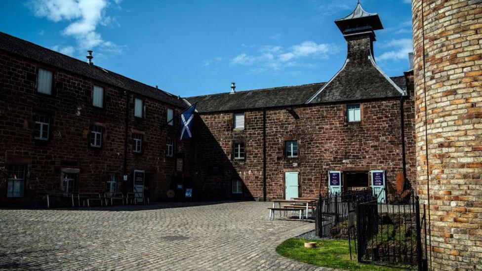 A traditional red brick building surrounds a courtyard. There are green shutters on the doors and windows, and a Saltire hanging from the roof. 