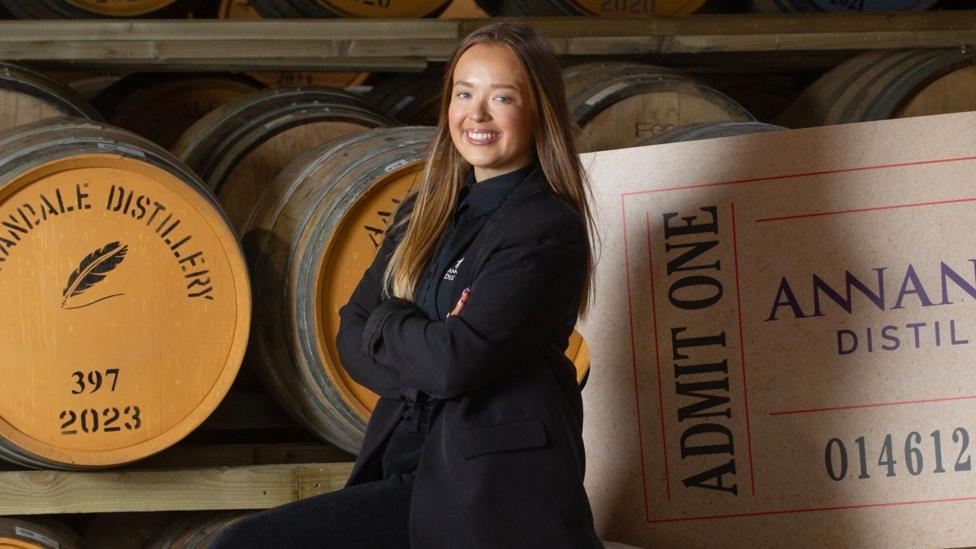 A young woman with long brown hair in dark clothing stands in front of whisky casks and a large ticket saying "Admit One"