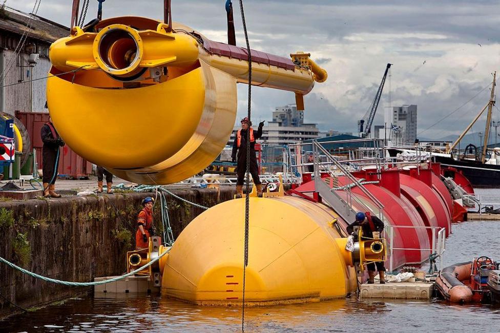 Large yellow marine energy device being lowered by crane into a harbor beside a floating structure, with several workers guiding the installation. Industrial waterfront buildings, vessels, and cranes are visible in the background under a cloudy sky.
